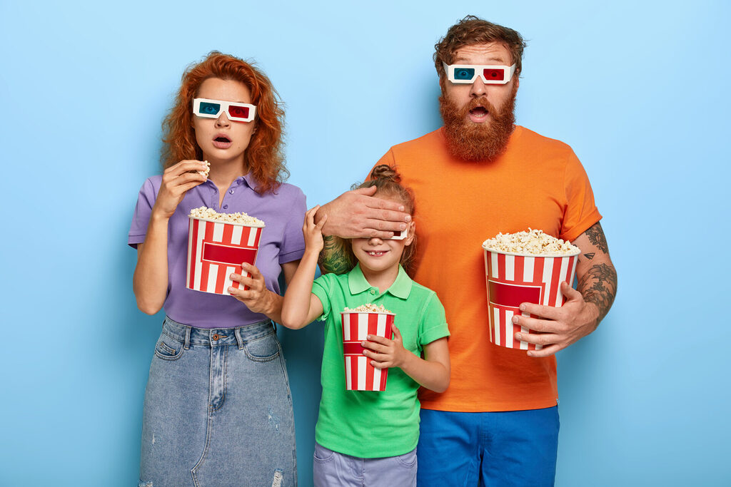 scared ginger family of three watch horror film, terrified by scarying scene, father covers eyes of small daughter not to see something awful, eat popcorn, go to cinema together during leisure time