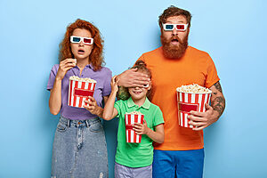 scared ginger family of three watch horror film, terrified by scarying scene, father covers eyes of small daughter not to see something awful, eat popcorn, go to cinema together during leisure time